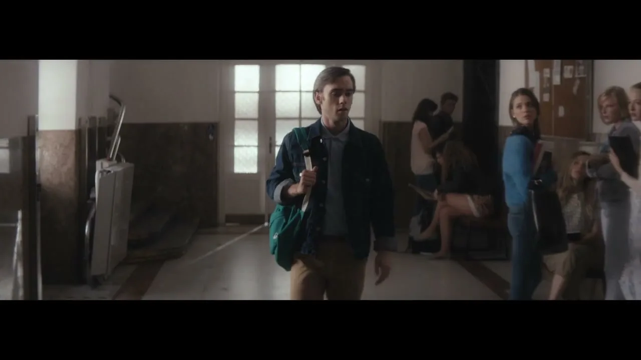A young man with a green bag walks through a school hallway, looking around as other students gather in the background.