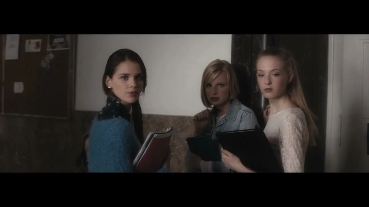 Three young women, dressed in casual attire, stand in a school hallway, looking intently towards the right side of the frame.