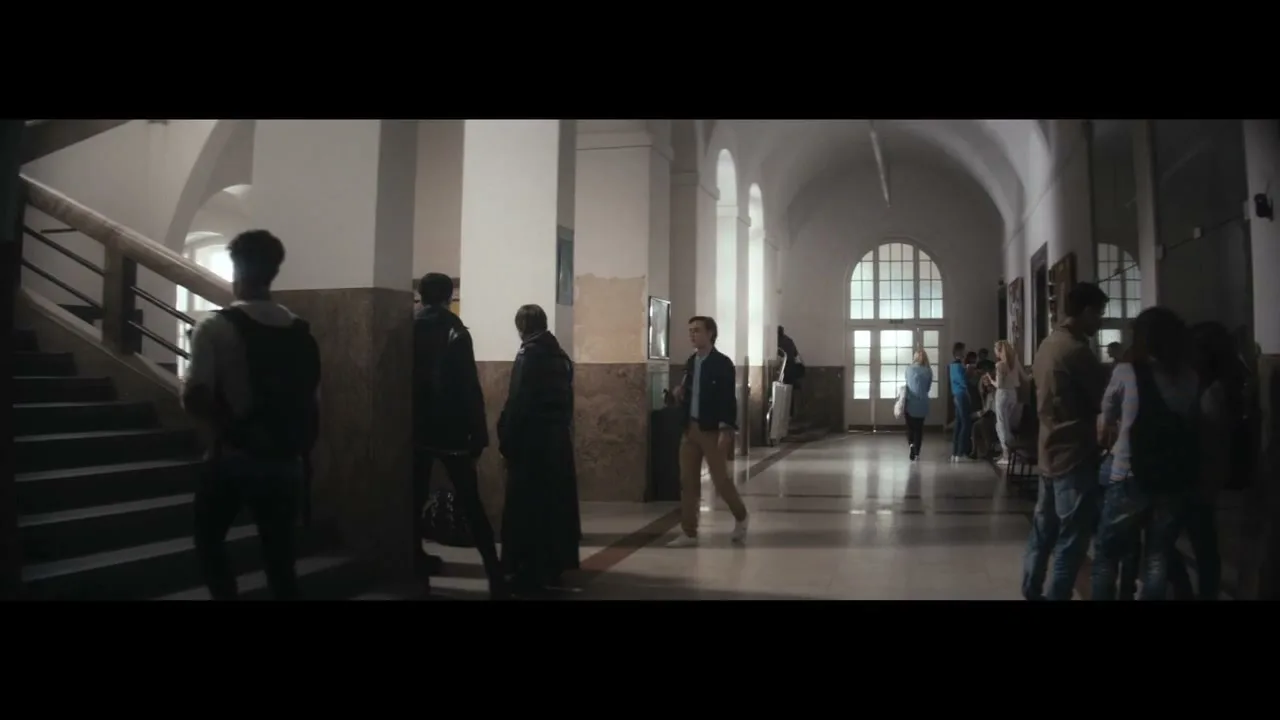 A young man in a blue shirt and backpack walks up a grand staircase in a school hallway, while other students mill about.