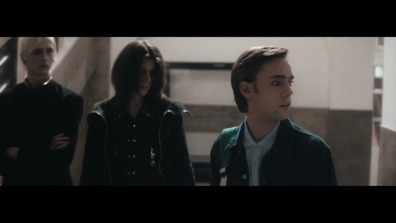 Three young men, two dressed in dark alternative fashion and one in a preppy style, stand in a school hallway, looking around with curious expressions.
