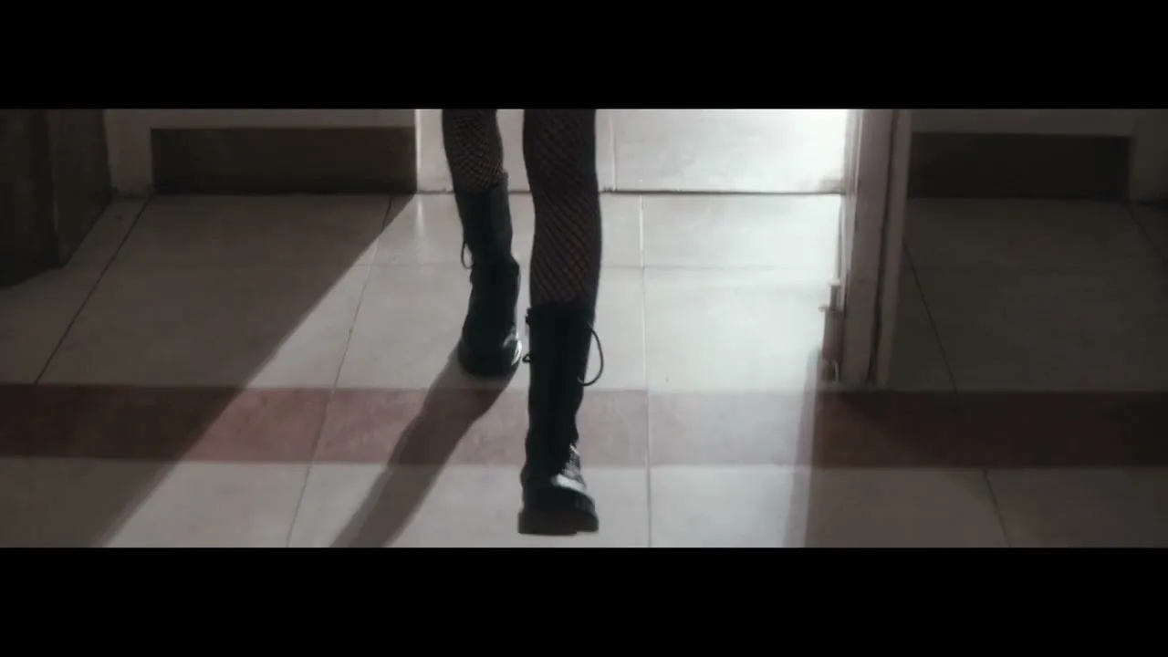 A woman in fishnet stockings and boots walks through a doorway into a dimly lit hallway, with sunlight casting long shadows on the tiled floor.
