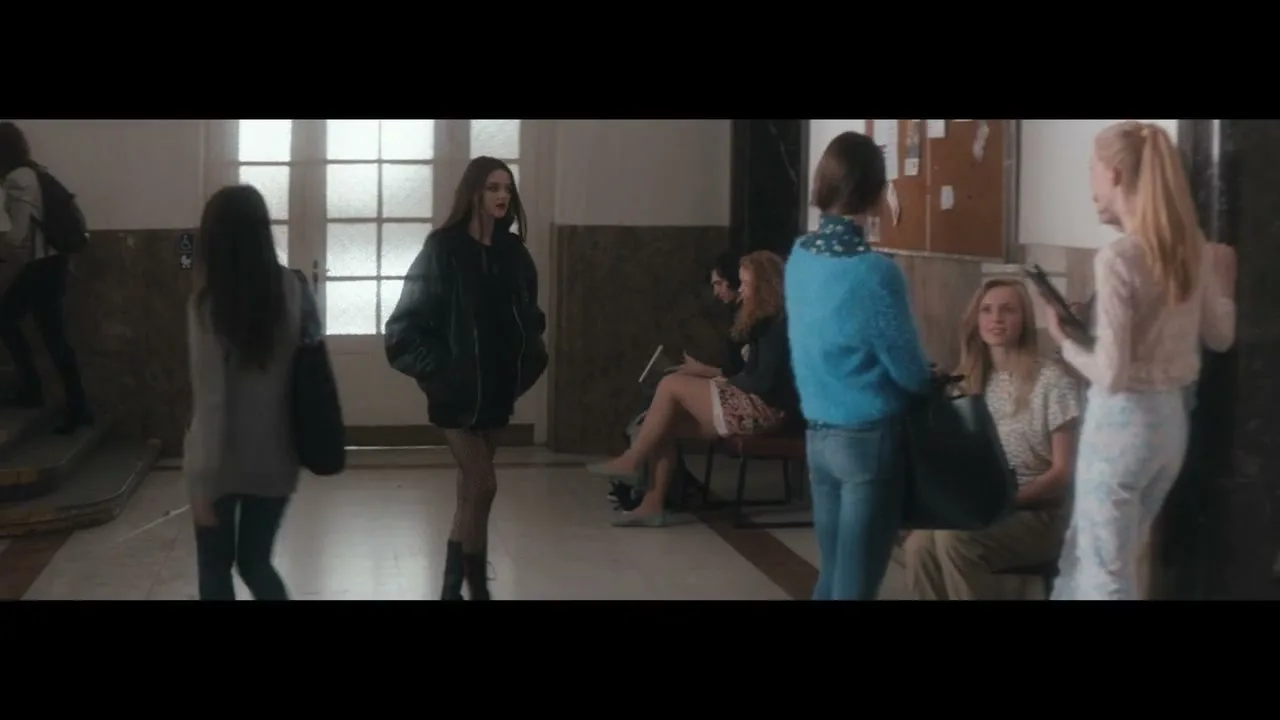 A young woman with long dark hair, wearing a black leather jacket and fishnet stockings, walks through a bright school hallway, looking around.