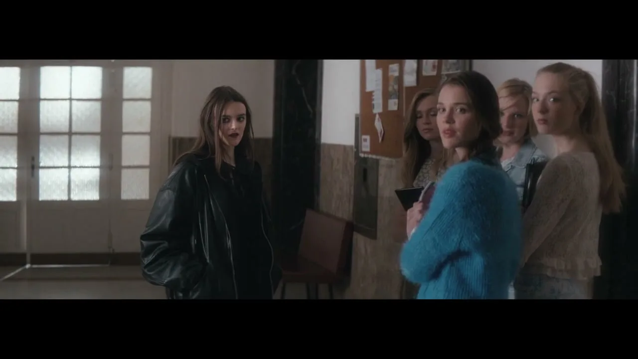 A young woman in a black leather jacket stands alone, looking at a group of four young women who are looking back at her in a school hallway.