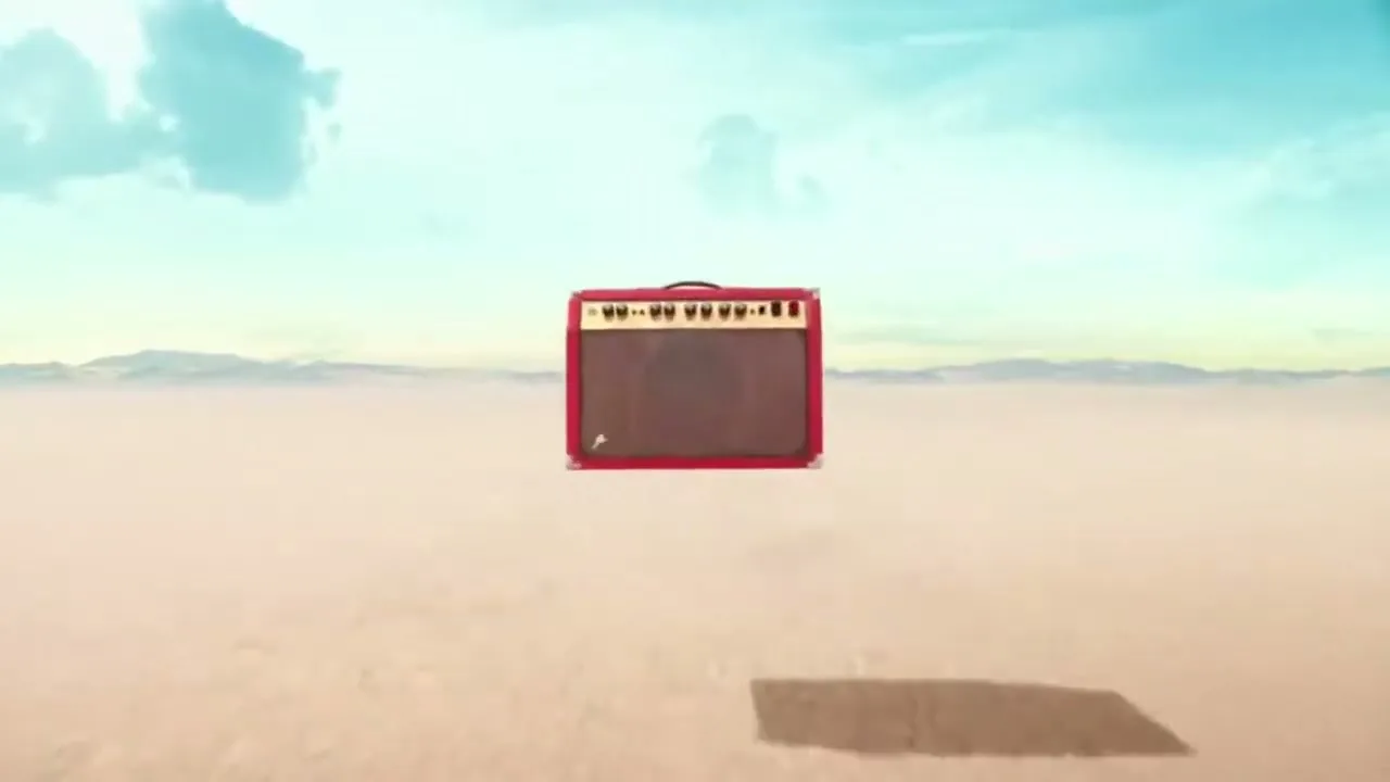 A red guitar amplifier sits in the middle of a vast, empty desert landscape under a bright blue sky, as the camera zooms in on it.