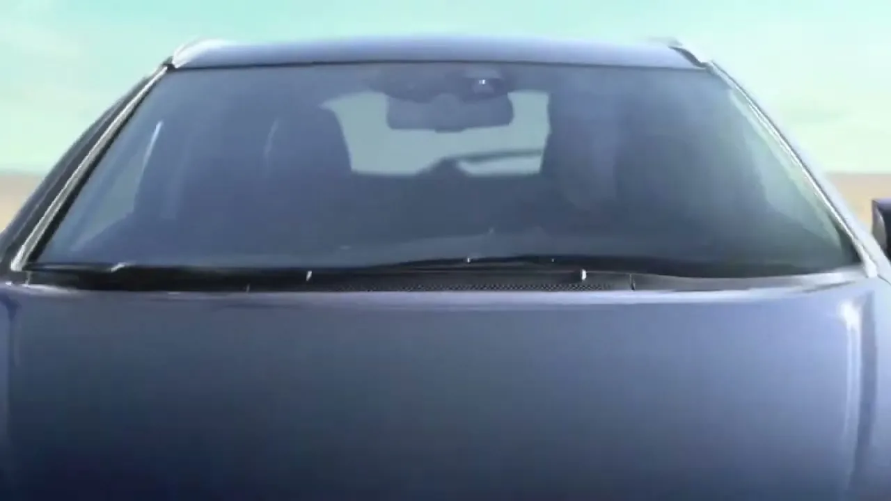 A dark blue Honda car is seen from the front, slowly moving forward on a light brown, sandy surface under a bright sky.