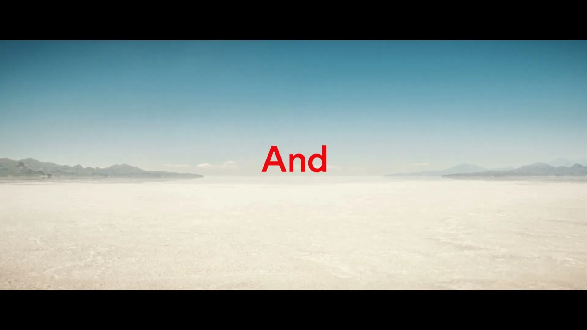 A wide shot of a vast, flat desert landscape under a clear blue sky, with mountains in the distance, as red text appears and disappears on screen.