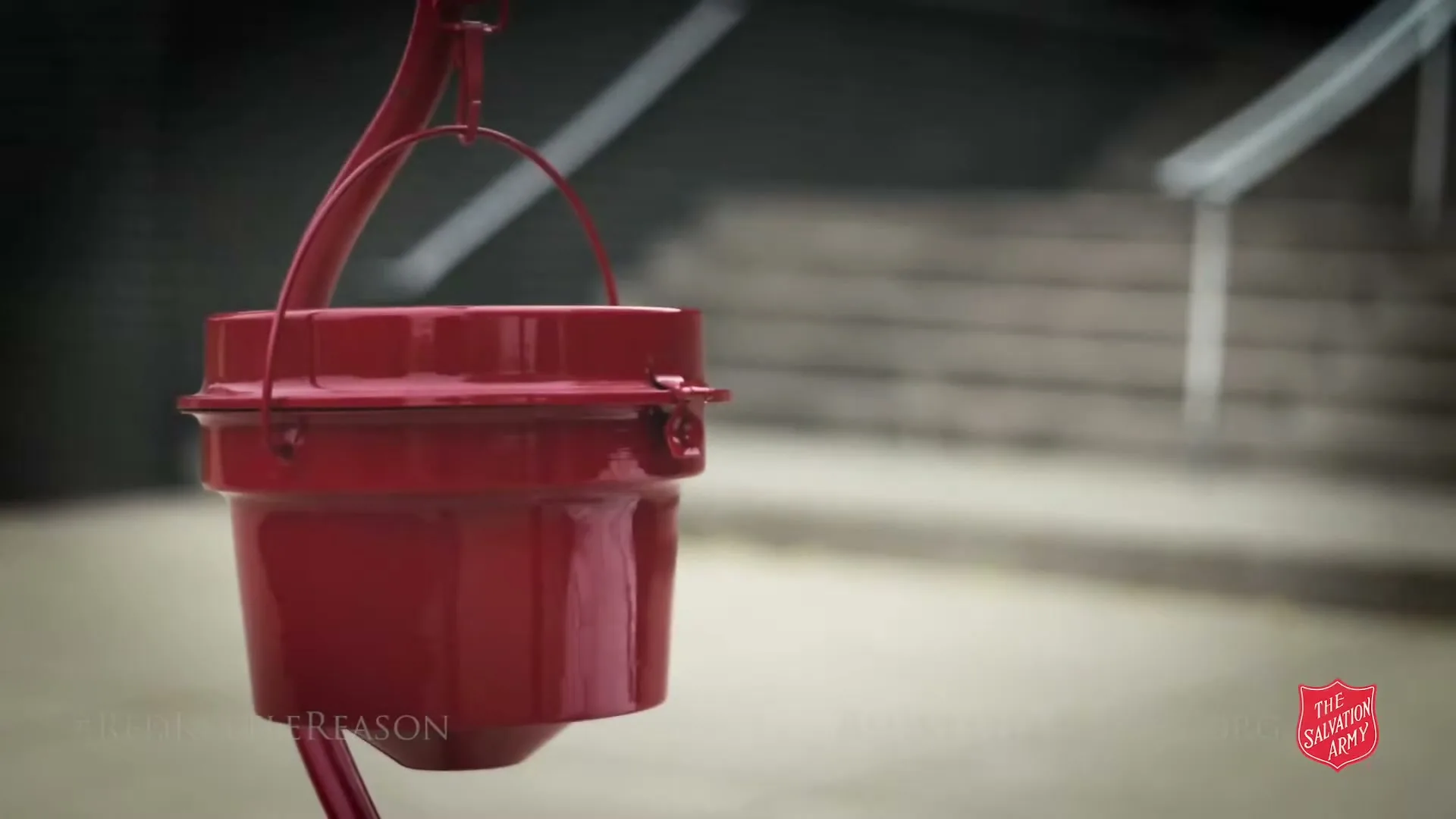 A red Salvation Army kettle hangs in the foreground, with blurred concrete steps and railings in the background, as text appears on screen.