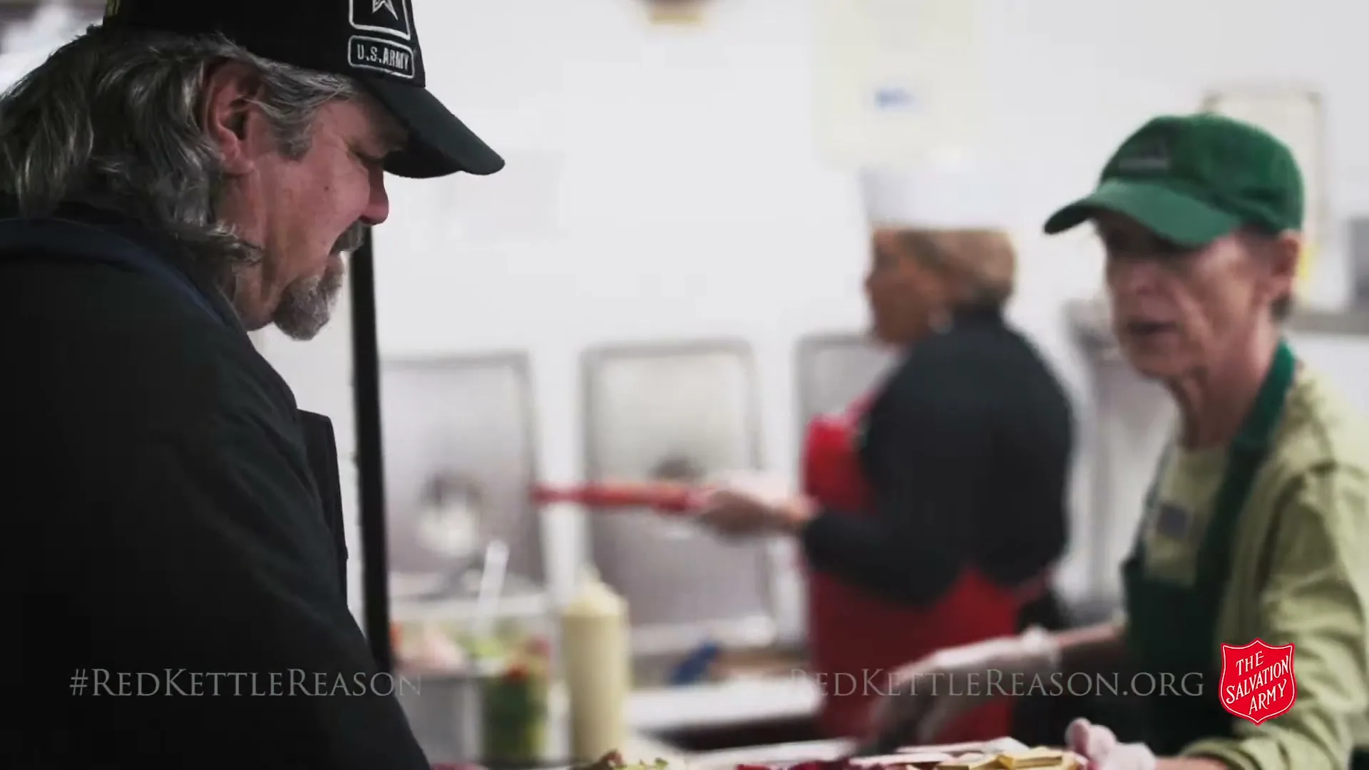 A man in a black U.S. Army cap and black jacket receives a plate of food from a server in a green apron and cap at a food service line in a brightly lit interior.