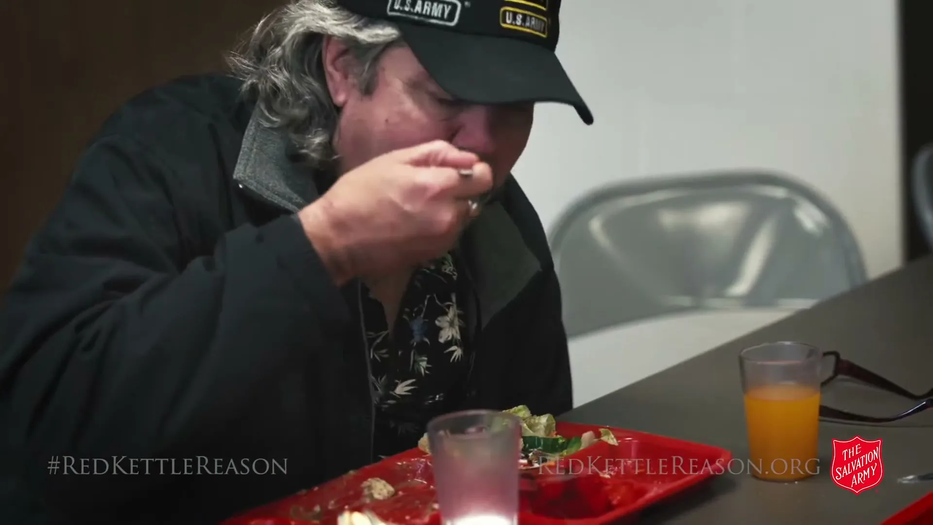 A man with a beard and a U.S. Army hat eats a meal from a red tray at a table in an interior setting.