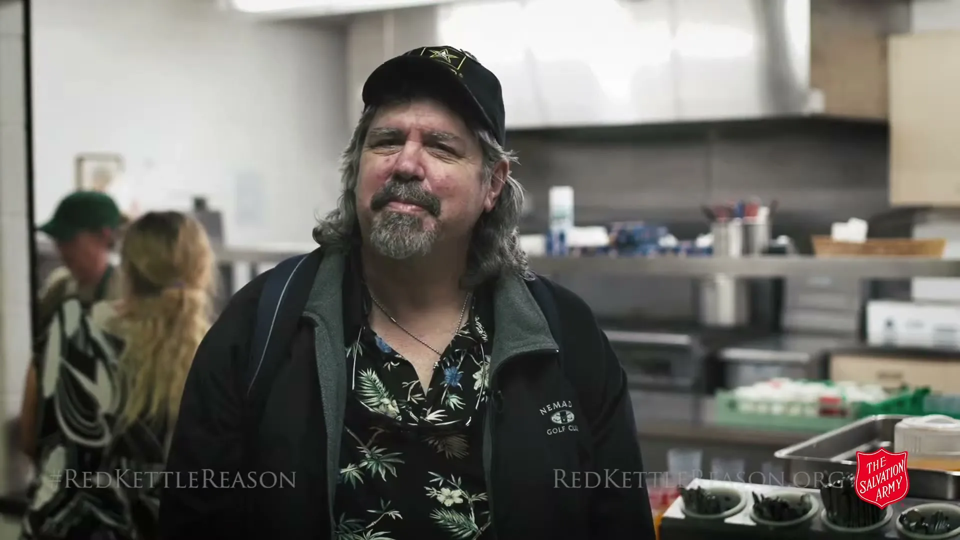 A man with a beard and long hair, wearing a black cap and a floral shirt, speaks to the camera in a kitchen.