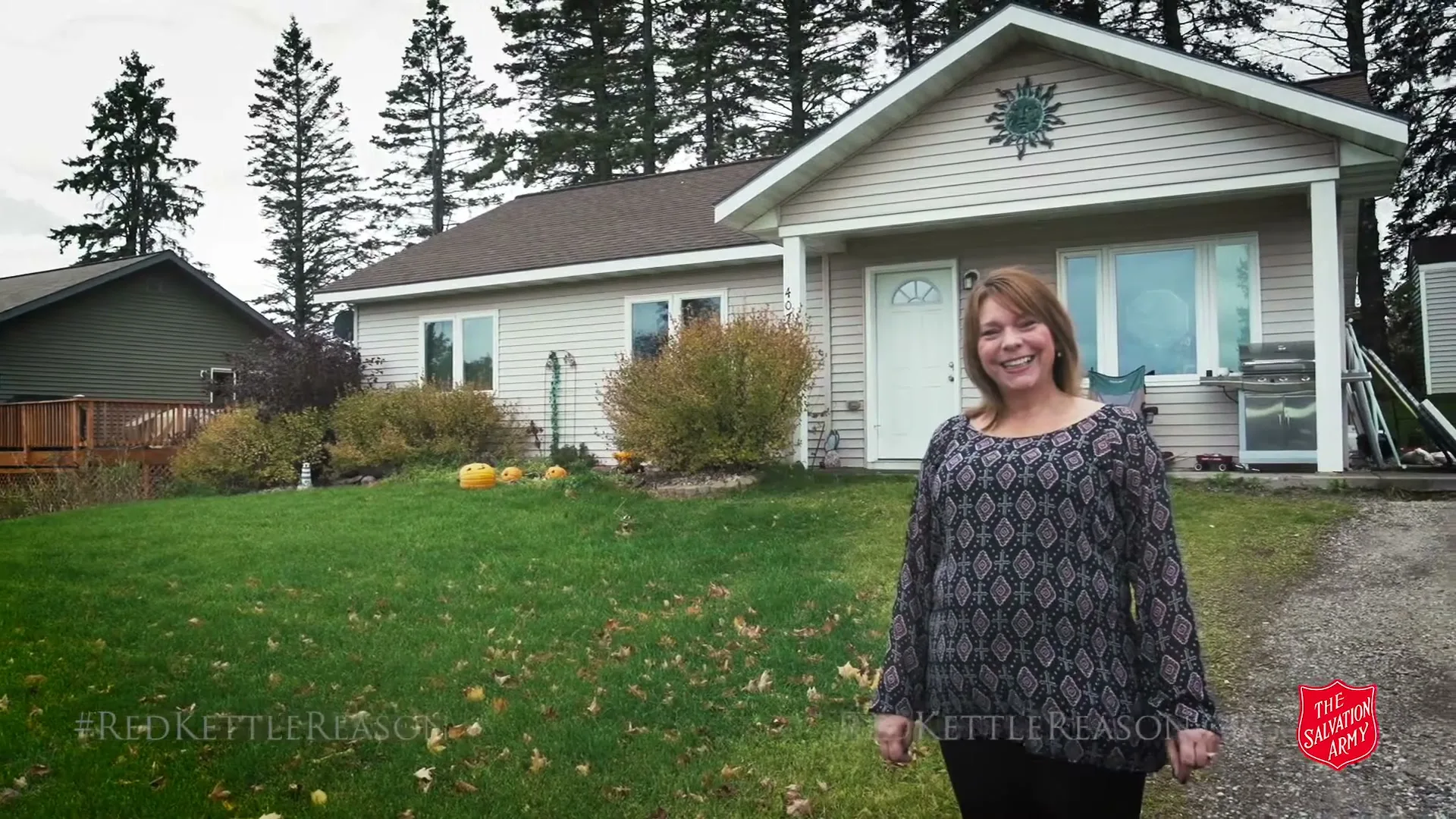 A smiling woman in her 40s stands in front of a small, light-colored house with a green lawn and pine trees in the background on an overcast day.