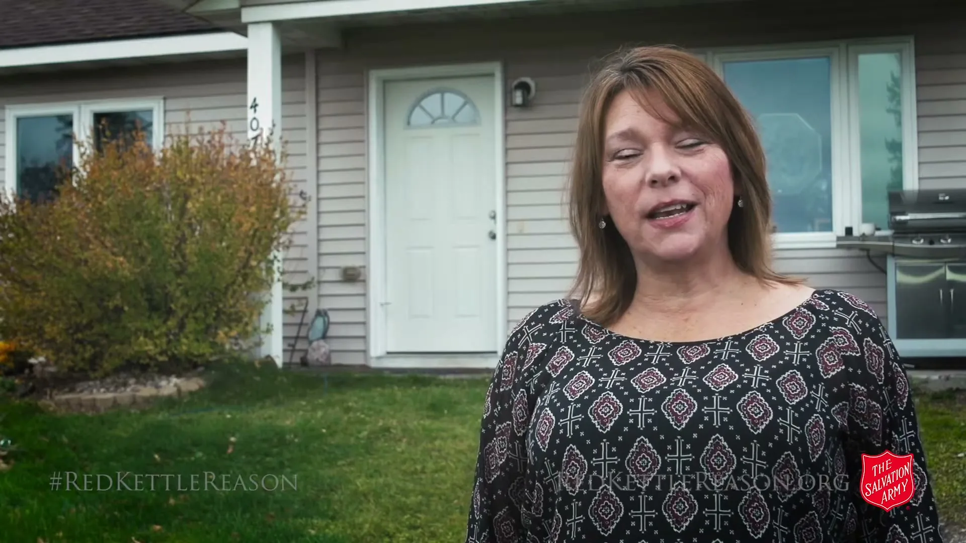 A middle-aged woman with brown hair and a patterned top speaks directly to the camera in front of a suburban house during the day.