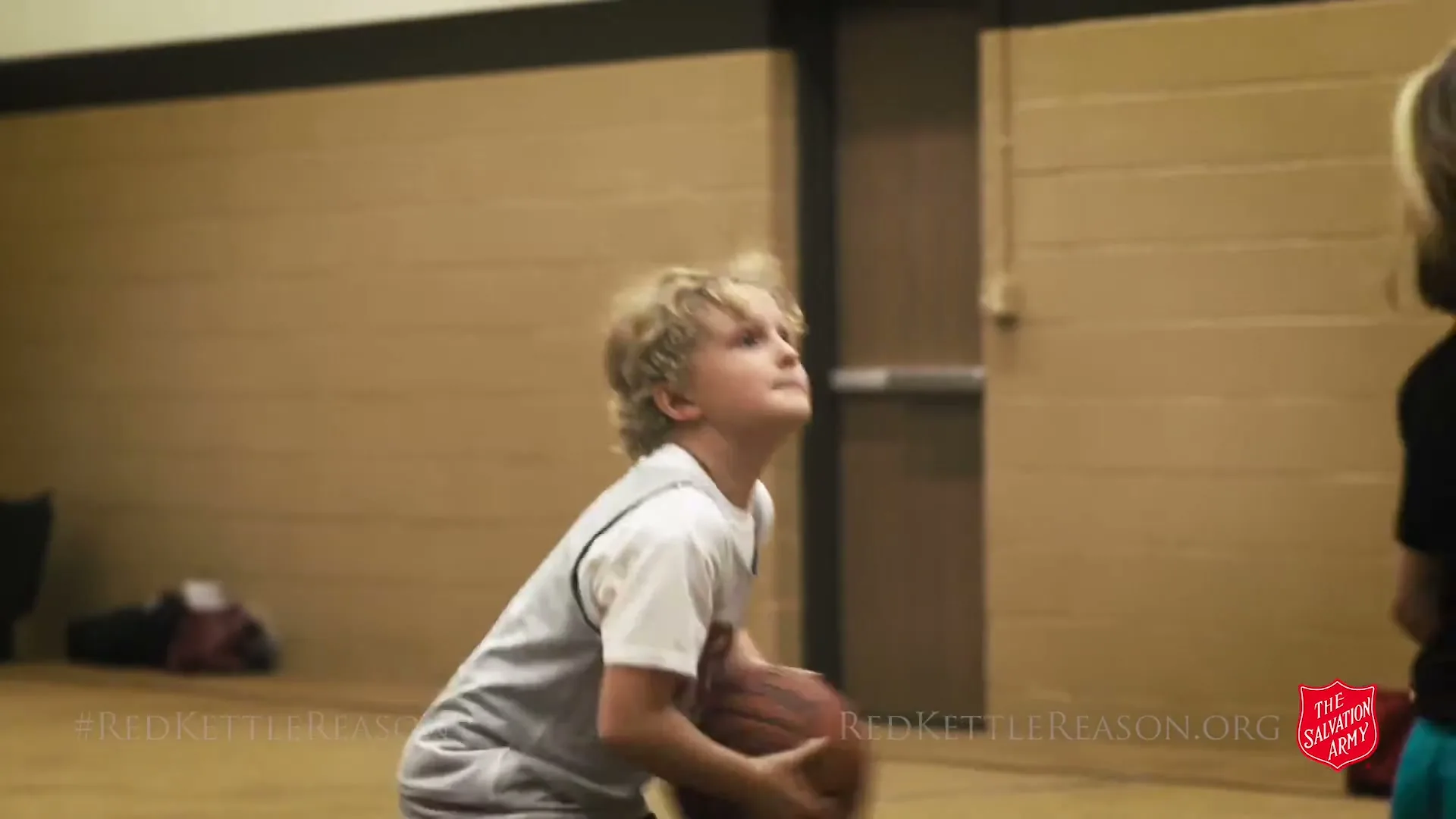 A young boy in a white and gray tank top shoots a basketball while a girl in a black shirt watches in a school gym.