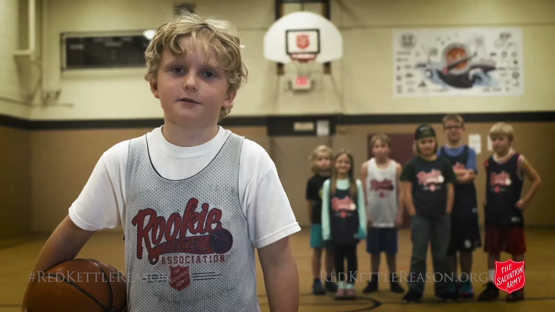 A young boy with blonde hair in a white t-shirt and basketball jersey holds a basketball and speaks to the camera, with other children in a line behind him on a basketball court.