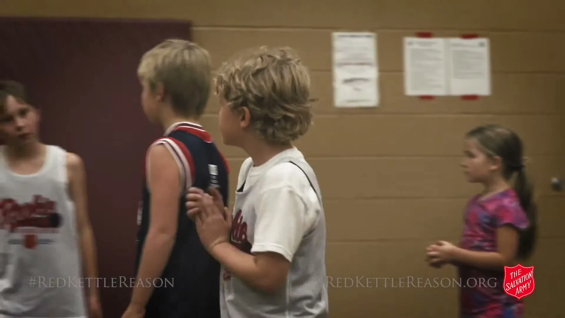 A young boy with curly blonde hair in a white and grey basketball jersey claps his hands while other children stand around him in a gym.