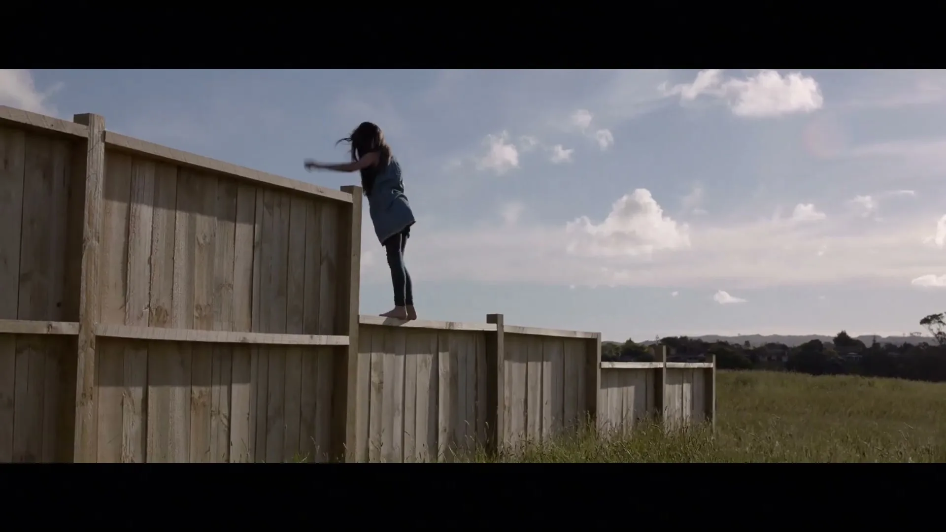 A young girl in a blue dress and dark leggings stands barefoot on a wooden fence, looking out over a grassy field under a cloudy sky.