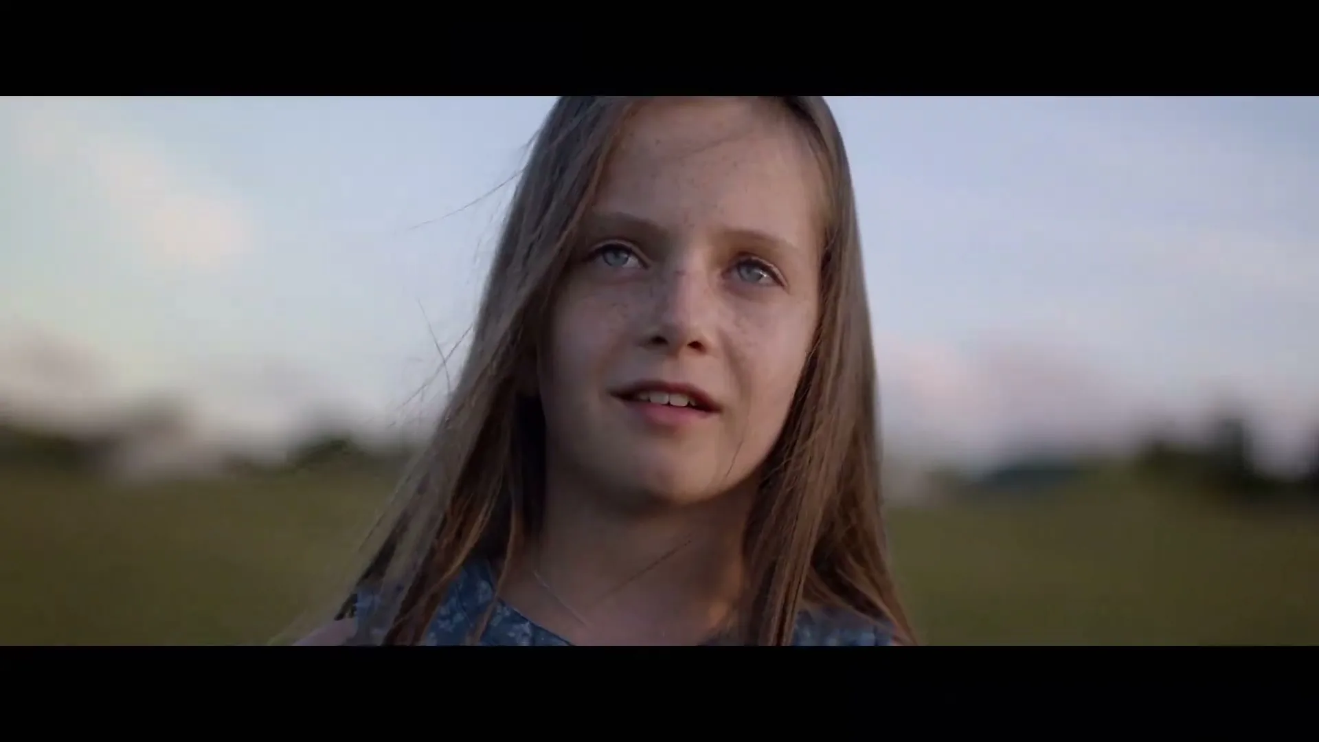 A young girl with long brown hair and freckles looks up with a slight smile in a field at dusk.