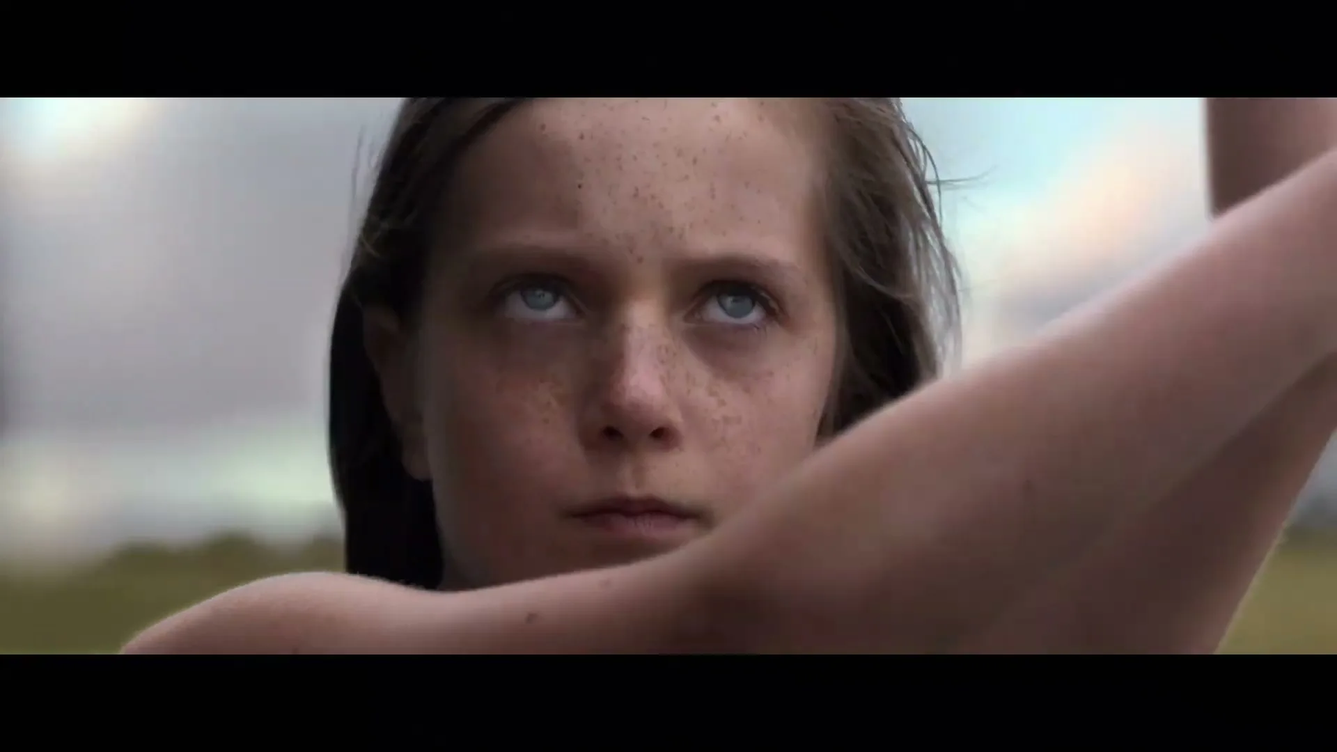 A young girl with freckles and blue eyes looks up intently, her arm raised, in a field under a cloudy sky.