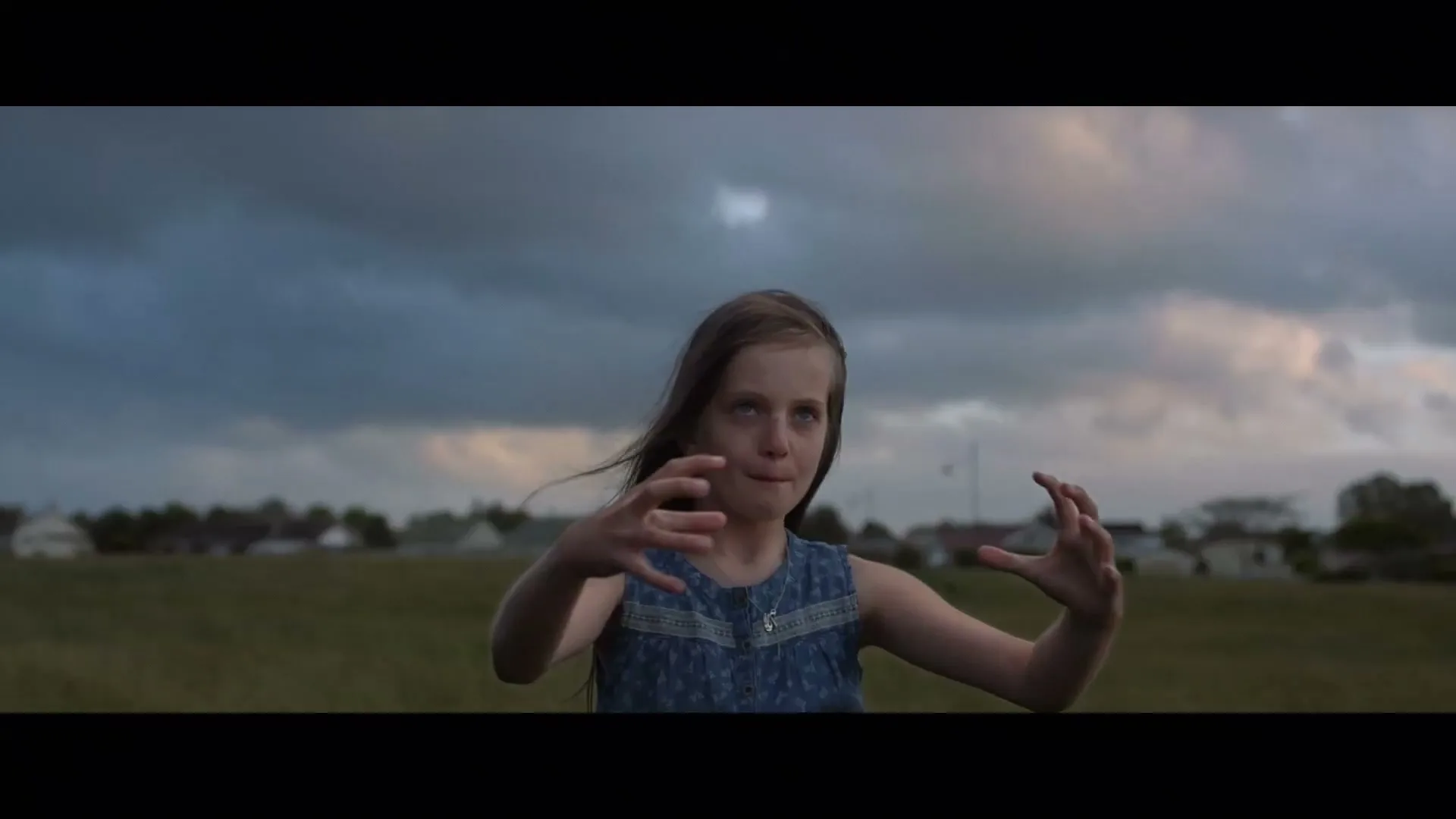 A young girl with long brown hair and a denim dress stands in a field, making a claw gesture with her hands under a dramatic sky.