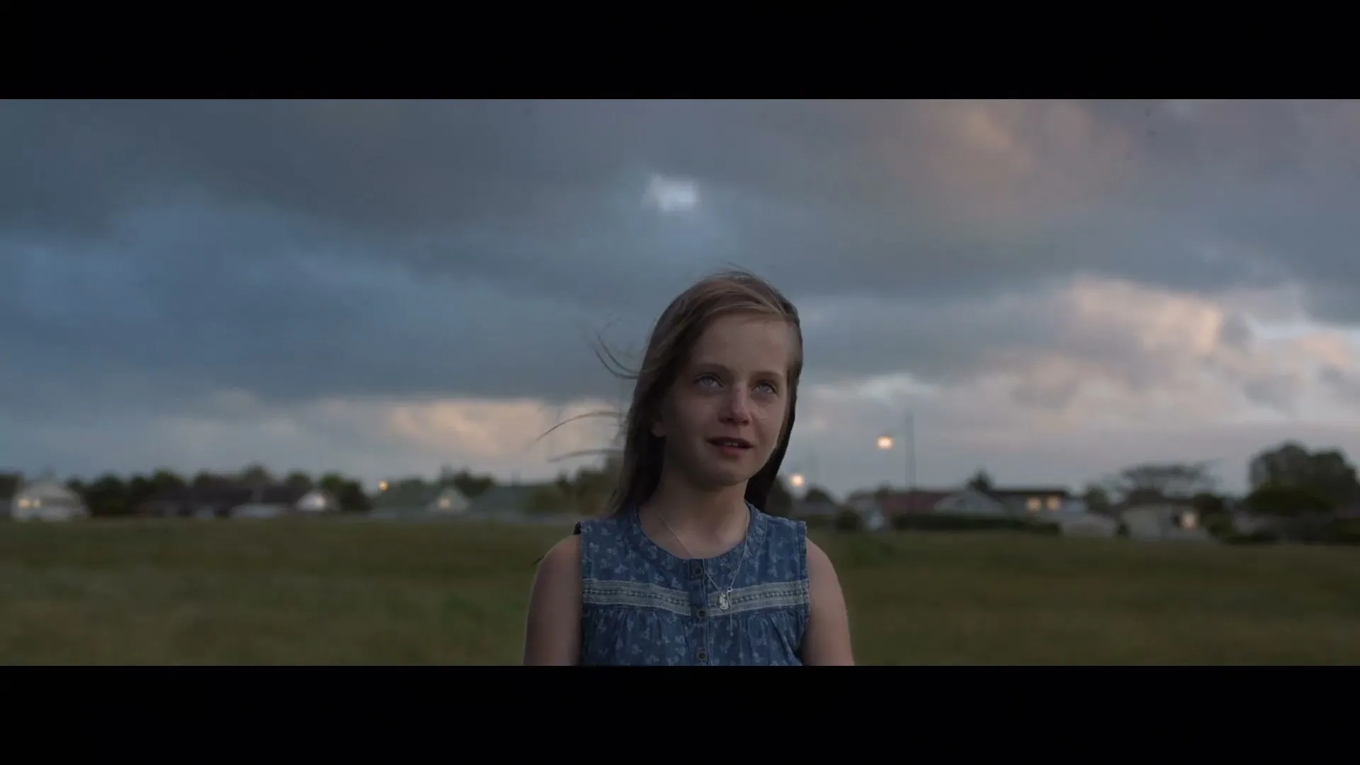 A young girl with long dark hair and a denim dress stands in a field, looking up with a slight smile as her hair blows in the wind under a cloudy sky.