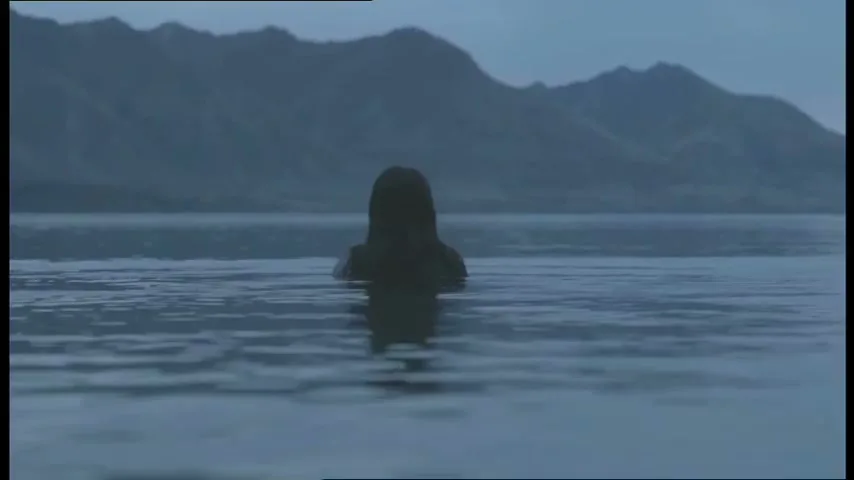 A woman with long dark hair is seen from behind, submerged in a lake with mountains in the background under a blue hour sky.