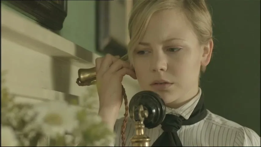 A young woman with blonde hair in a period costume listens intently on an old-fashioned telephone in an interior setting.