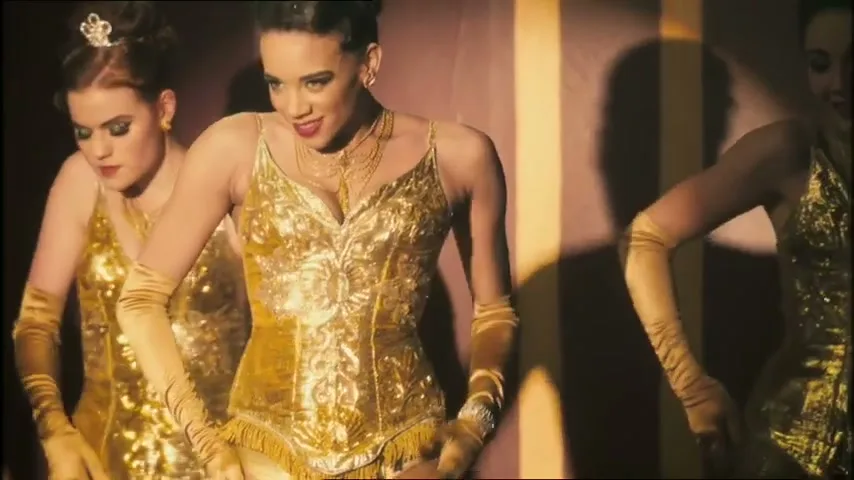 Three female dancers in golden burlesque costumes perform on a dimly lit stage with dramatic shadows.