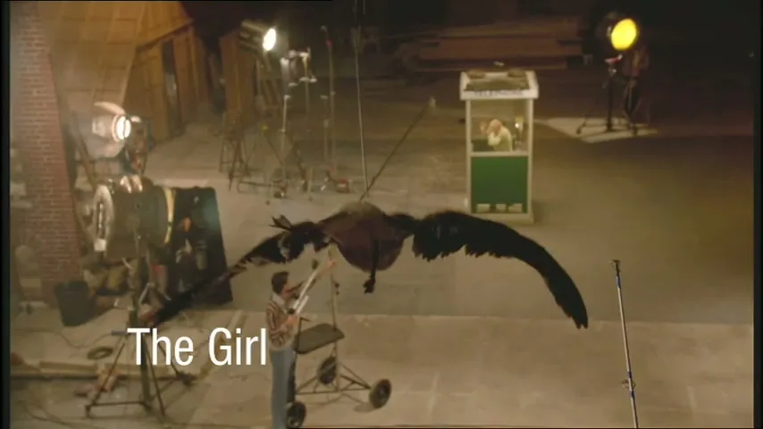 A large, dark bird puppet with wide wings is being manipulated by a male puppeteer in a dimly lit studio with various film equipment.
