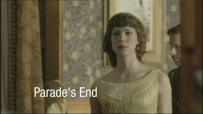 A woman in a gold dress and headpiece looks forward, while a man in uniform stands behind her in a period room.