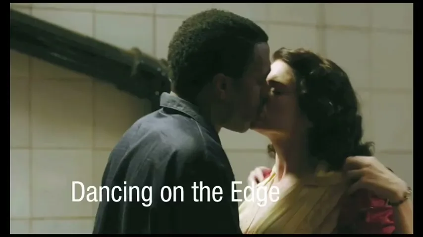 A Black man in a dark shirt kisses a white woman in a red top and yellow dress in an interior setting.