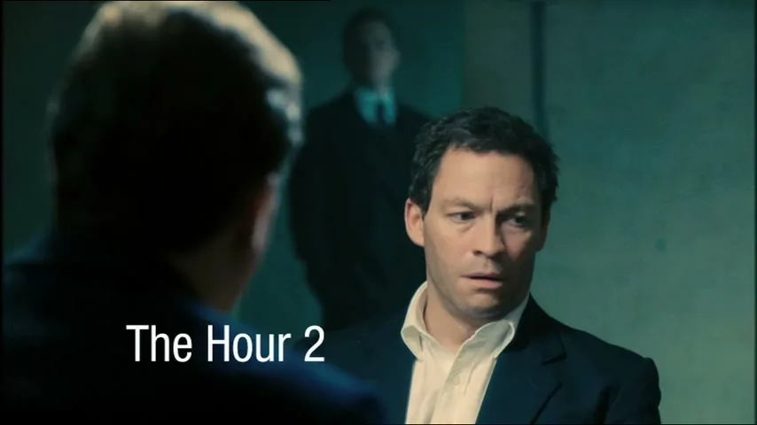 A man in a suit looks concerned while another man's back is to the camera, with a third man blurred in the background, in an interior setting.
