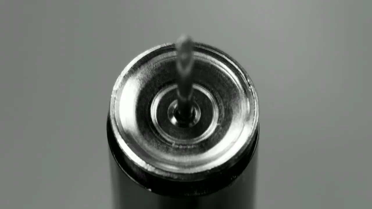 A close-up shot shows a small, sharp metal pin being released from a cylindrical object, rising upwards against a grey background.