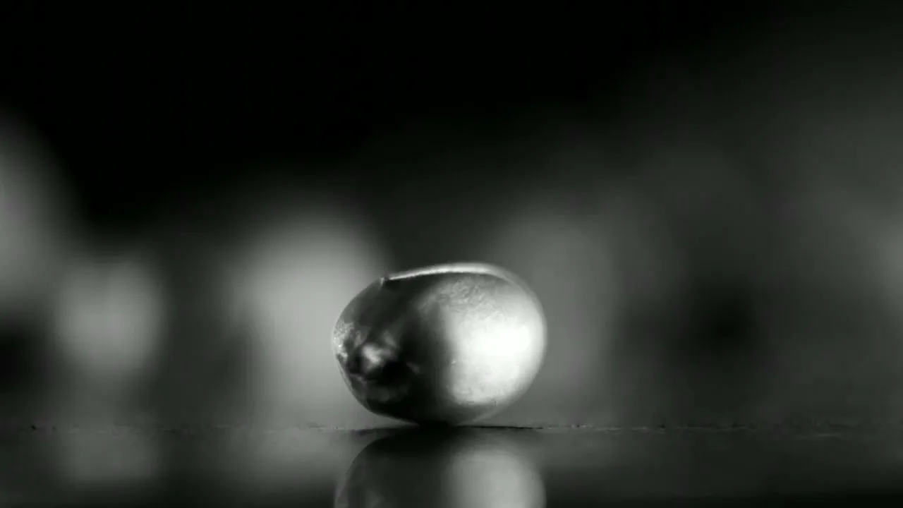 A single, small, metallic ball rests on a reflective surface in a dark, minimalist setting, highlighted by a strong light source.