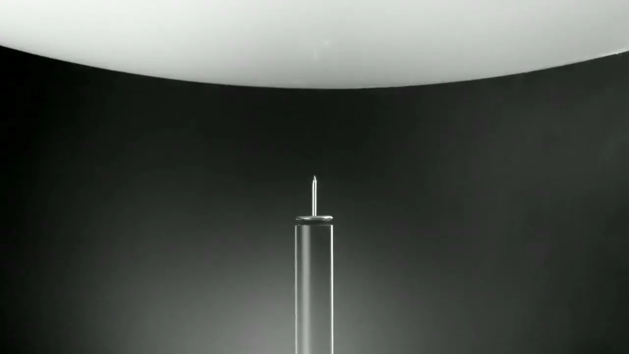 A white balloon slowly descends onto a sharp nail, poised to pop, in a studio void.