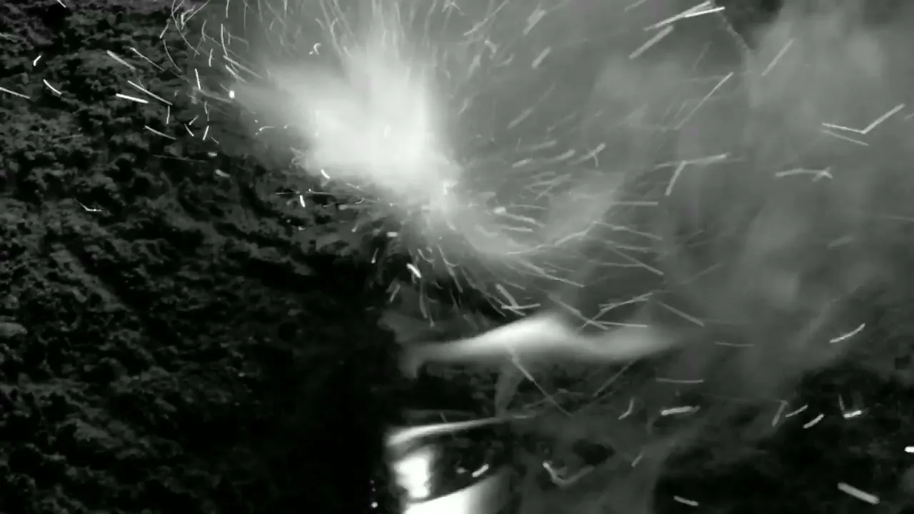 A black and white, high-angle shot shows a small firework exploding, emitting sparks and smoke against a dark, textured background.