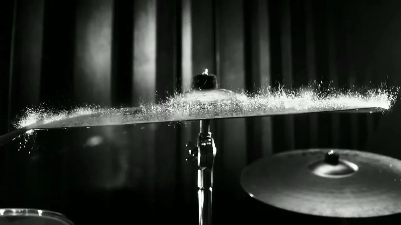A black and white shot of a drum cymbal with water droplets on it, vibrating and splashing water upwards in a studio.