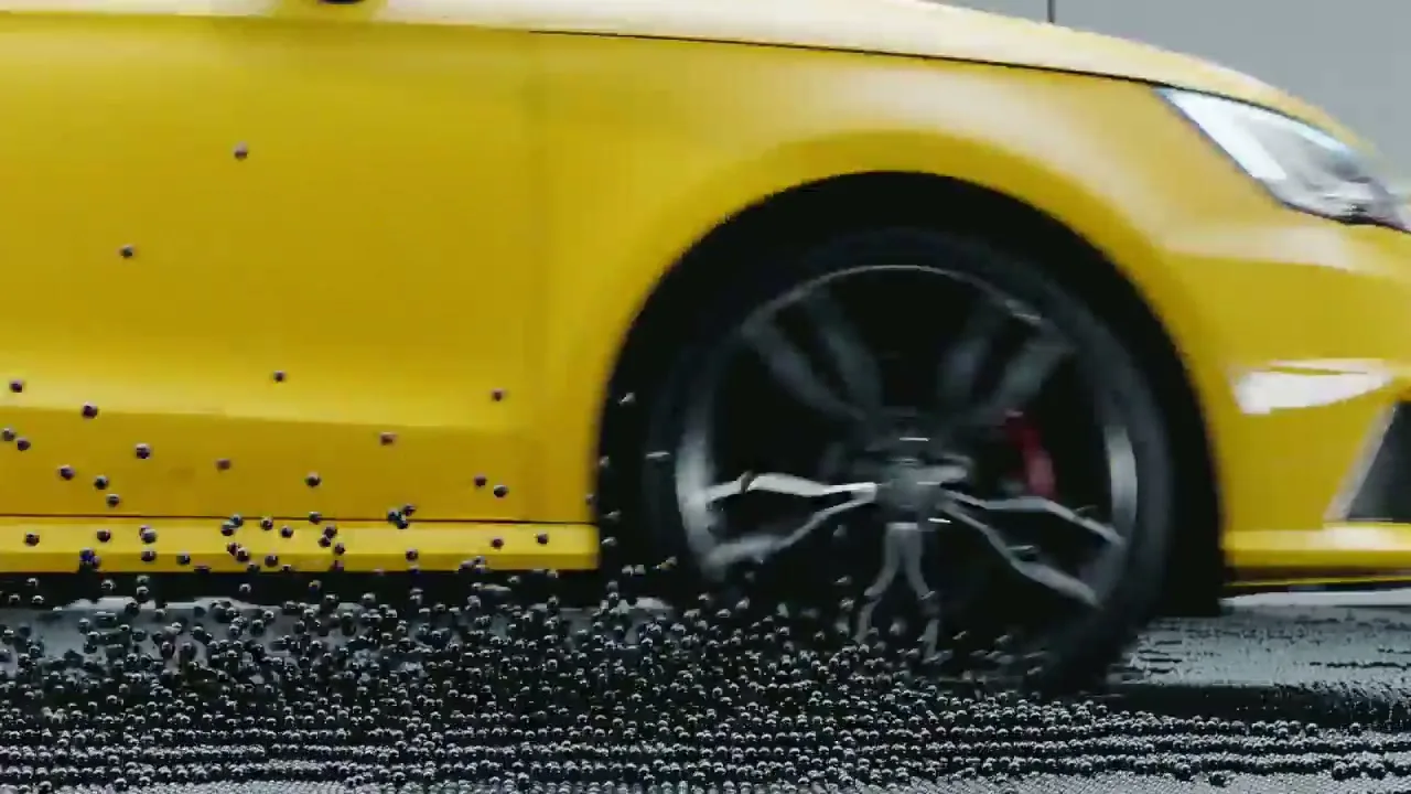A yellow Audi car drives across a surface of small black balls, which scatter as the car moves.