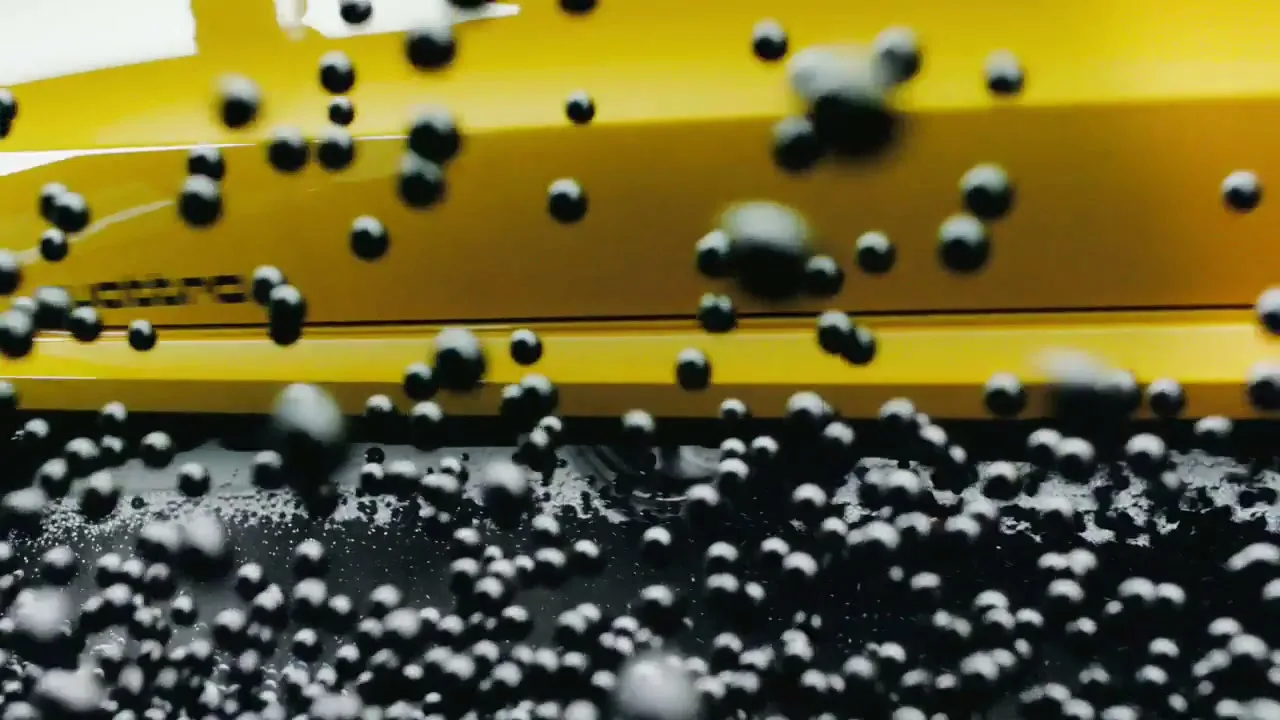 A yellow Audi car is seen from a low angle as small black beads bounce off its side and wheel, creating a dynamic visual.
