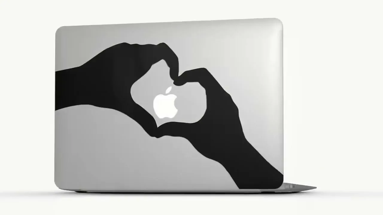 A silver MacBook laptop with a black sticker of two hands forming a heart around the Apple logo rotates on a white background.