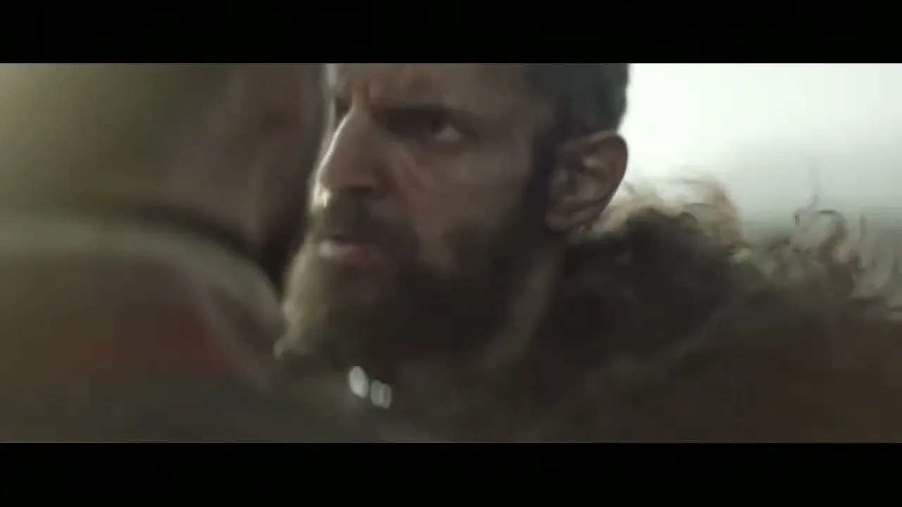 A bearded man with a fur cloak looks intensely at another person, his face showing determination in a misty, outdoor setting.