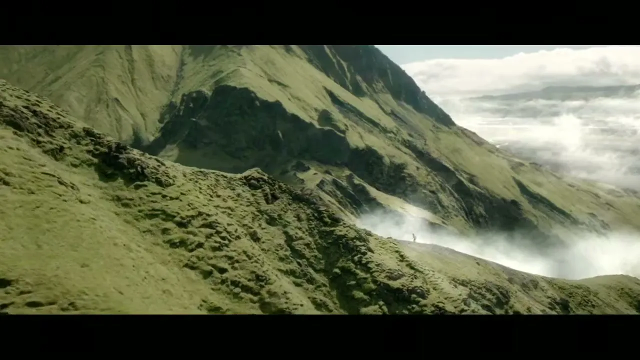 A wide shot of a vast, green mountain range with a small figure standing on a ridge, surrounded by mist and clouds.