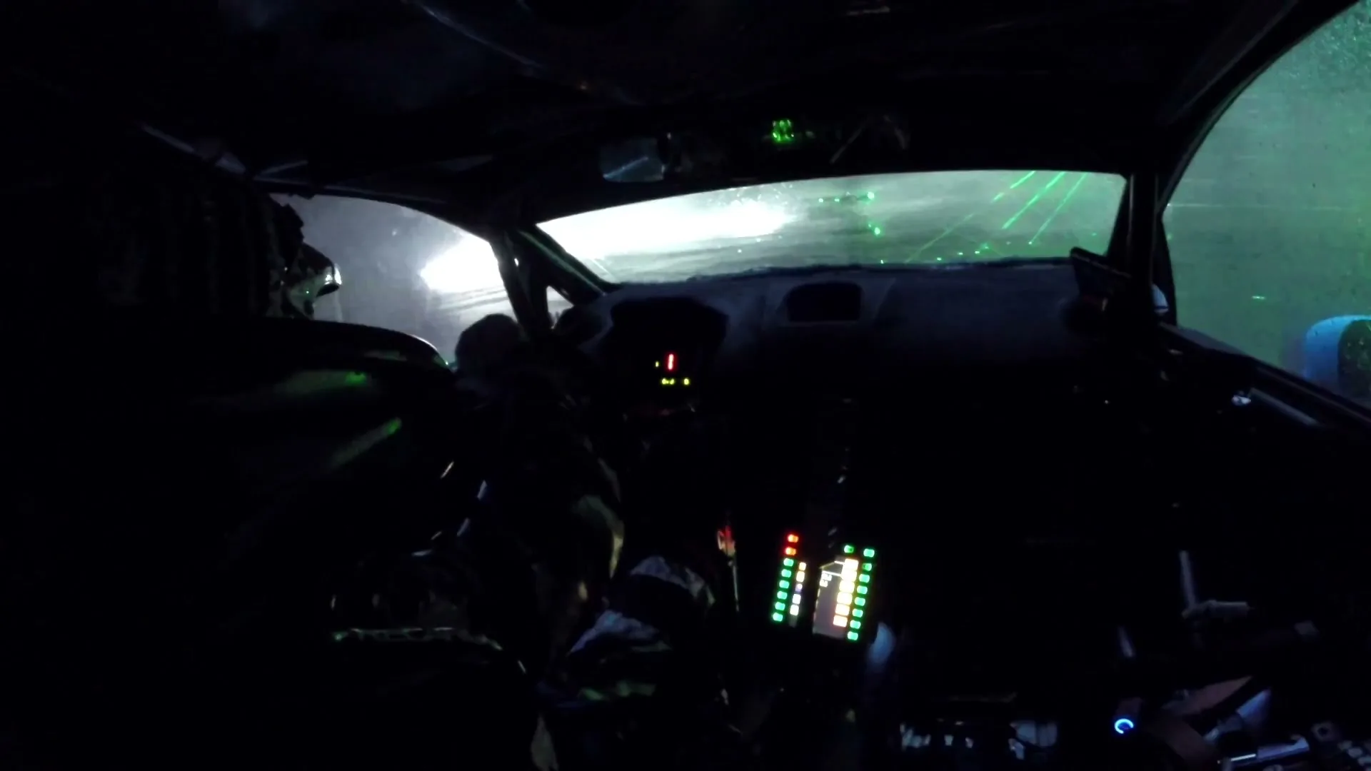A dark interior shot of a race car with a driver and co-driver, as green laser lights illuminate the track ahead, simulating a night race.