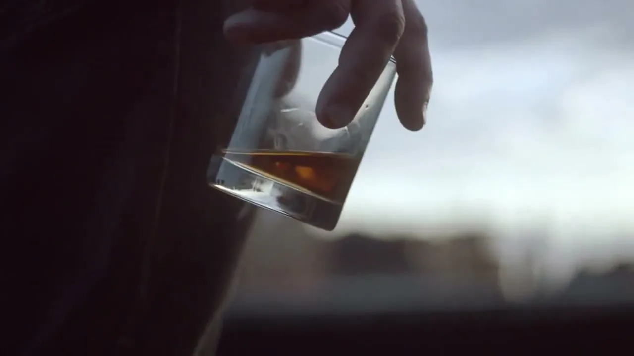 A person's hand holds a glass of amber liquid, possibly whiskey, against a blurred outdoor background with a cloudy sky.