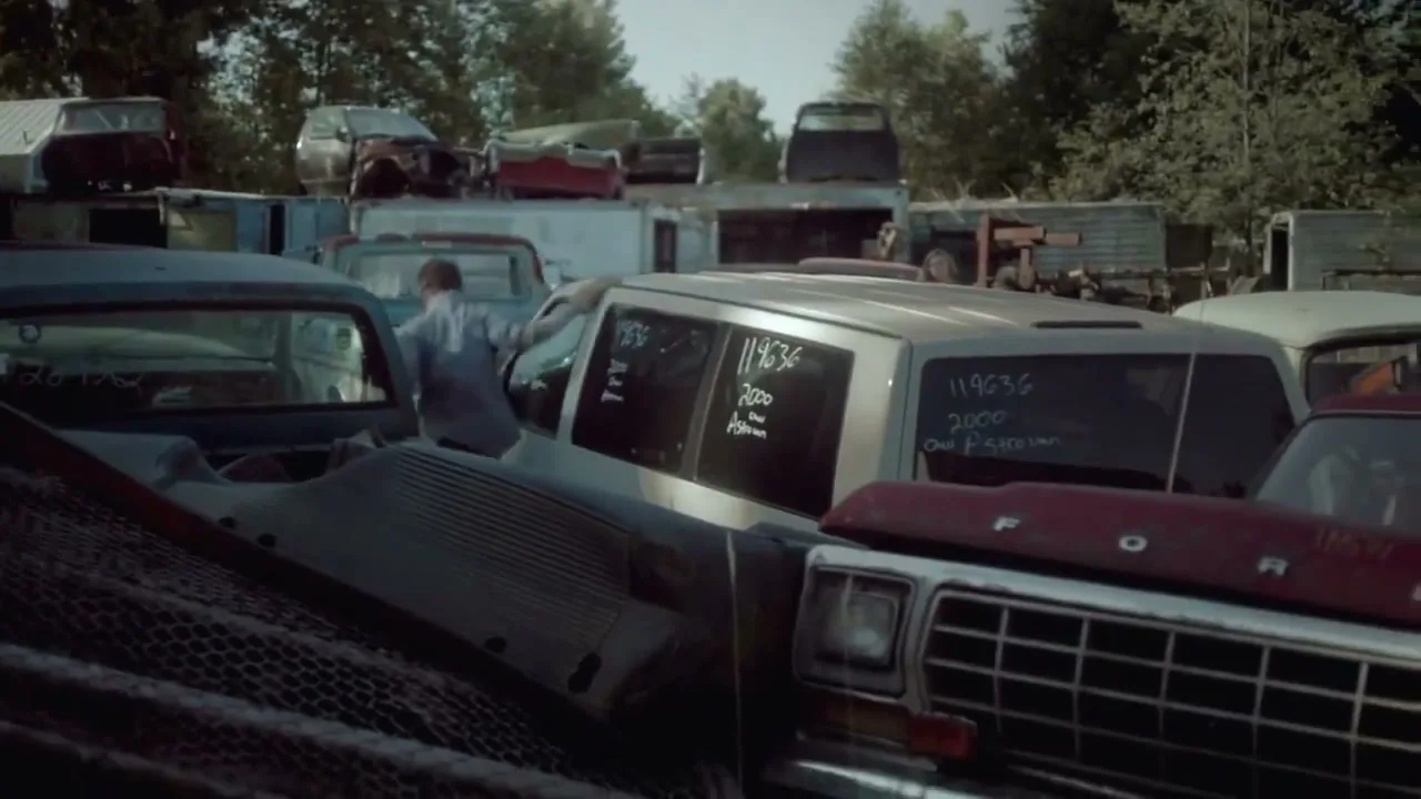 A young man in a light blue shirt and dark pants climbs over old, rusted cars in a junkyard on a sunny day.