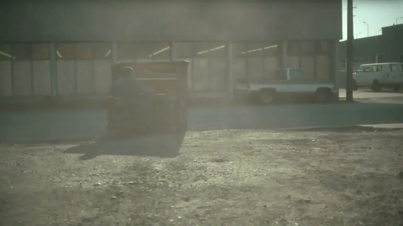 A man in a blue shirt and jeans plays a piano on a dusty lot in front of a building with large windows and a parked pickup truck.