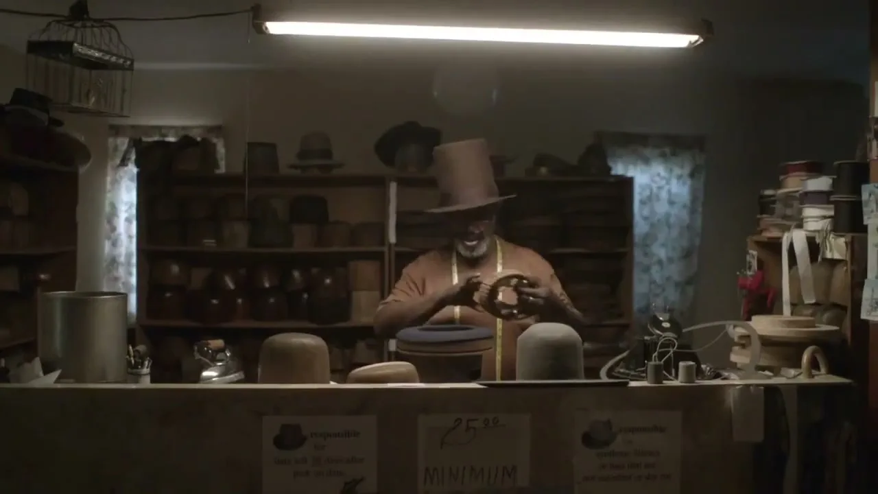 A man in his 60s, wearing a top hat and an orange shirt, works on a hat behind a counter in a dimly lit hat shop.