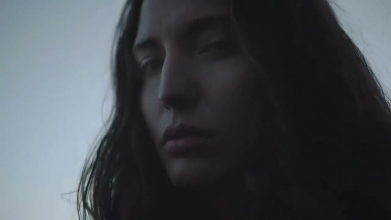 A young woman with long dark hair looks intently at the camera, her face partially obscured by shadow against a muted sky.