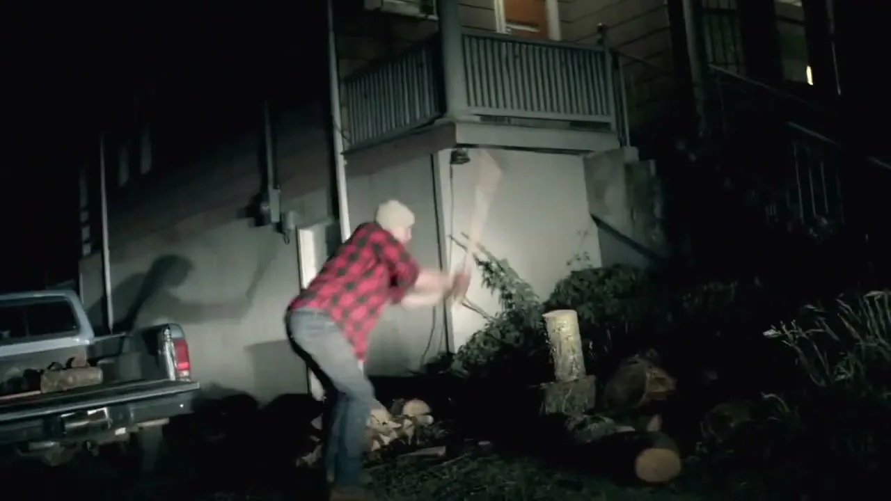 A man in a red plaid shirt chops wood with an axe at night, next to a house and a pickup truck.