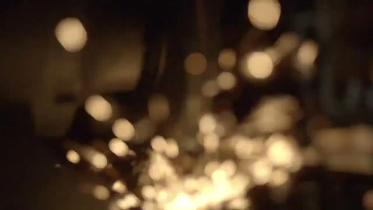 A blurred shot shows bright sparks flying upwards from an unseen source, with dark, indistinct shapes in the background.
