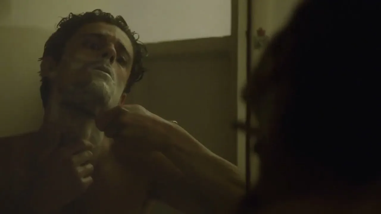 A man with dark, curly hair and shaving cream on his face shaves in a mirror in a dimly lit bathroom.